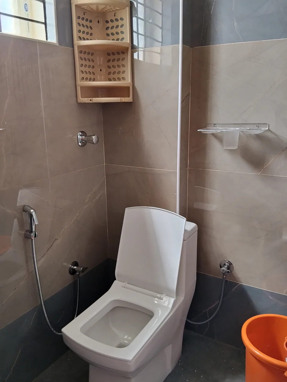 Tiled washroom with a toilet, shower fittings and an orange bucket.